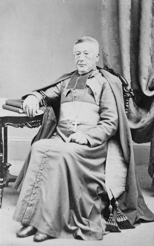 Black and white photograph of an elderly man wearing the liturgical garments of a bishop. He does not wear the cap. He sits on a chair, his right arm resting on a table, his hand resting on books. He wears a serious expression.
