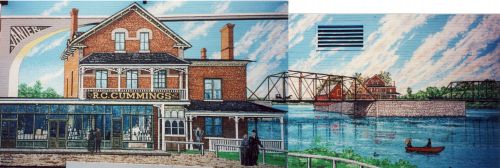 Color photograph of a mural depicting near and far views of the “R.C. Cummings” commercial building in red brick. The building, located on an island in the center of a river, is reached by a metal bridge.