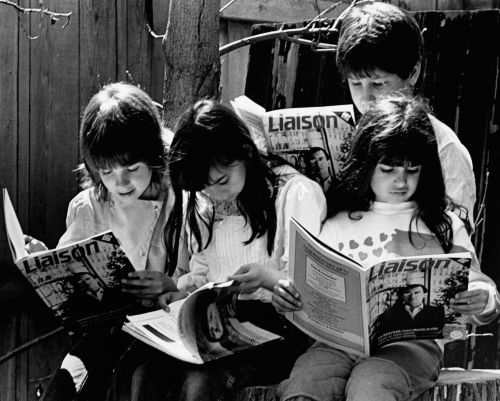 Black and white photograph of four children, each one reading a copy of Liaison magazine, which they hold in their hands. They are outside. Two girls and a boy sit in a row on log stumps, with another boy standing behind them.