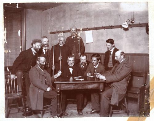 Black and white photograph of a group of men around a table. Five men are seated. Five others, including one cleric, are standing, some holding billiard cues. Two of the seated men hold cards in their hands. The photograph is slightly damaged.