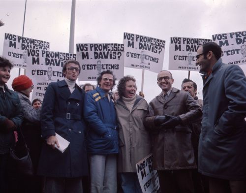 Photographie en couleur de quatre hommes et d’une femme d’âge mûr, à l’avant d’un groupe de manifestants. Ils sourient et tiennent des pancartes où il est écrit : « Vos promesses, M. Davis ? C’est l’temps. On est 600 000 ! »