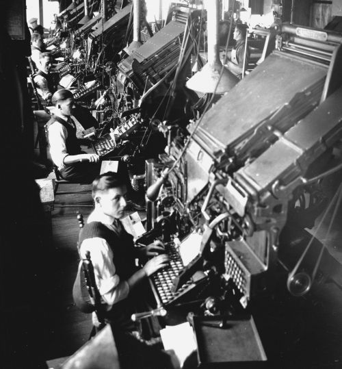 Black and white photograph of five men in a shirt, vest and tie in an industrial workshop. They sit in front of imposing machines of composition, with keyboard.