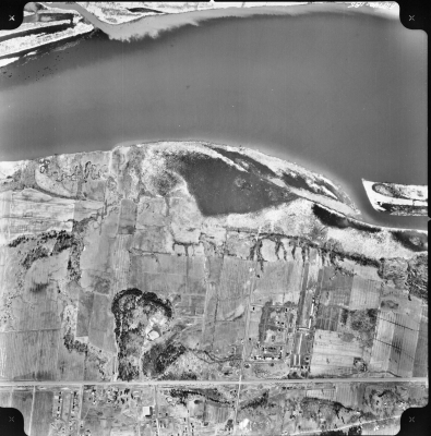Photographie aérienne en noir et blanc d’un paysage campagnard où se trouve, au nord (au haut de l'image), une rivière. Quelques habitations sont regroupées dans la portion du territoire au sud (au bas de l'image), le long de deux routes principales.