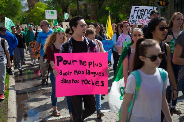 Colour photograph of a group of demonstrators out on the streets. Many of them are sporting green and white, colours of the Franco-Ontarian flag. One of them holds a sign that reads: “Pour ne plus avoir nos langues dans nos poches” (To no longer have to bite our tongues).