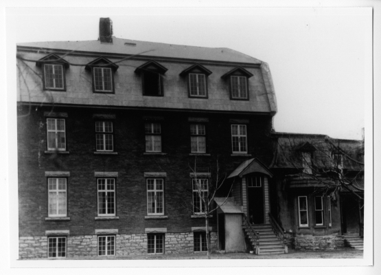 Photographie en noir et blanc d’un édifice en brique de trois étages, à toit mansardé. Un petit escalier mène vers un portique, qui fait office de porte d’entrée. Une maison attenante de plus petite taille le borde à droite.
