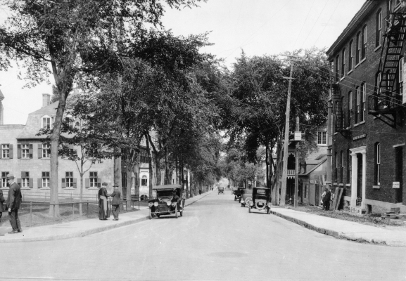 Black and white photograph of a busy street lined with trees, also many cars and pedestrians. Residences line one side of the street, with an institutional building situated on the opposite side. Men,  including a man of the cloth, talk on the sidewalk.