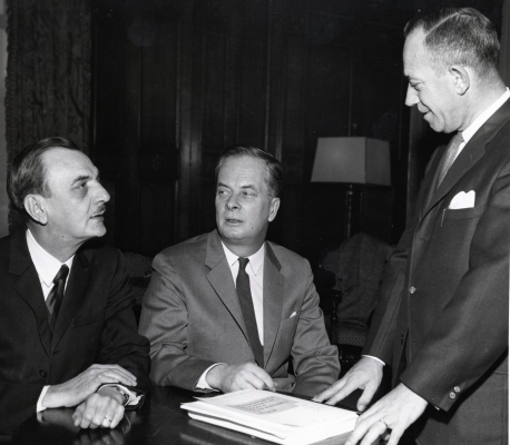 Black and white photograph of three older men in suits and ties. Two of the men sit side by side at a desk with a stack of papers in front of them. The third man stands, facing them. One man is seen in full-face view, the others in profile.