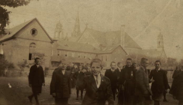Sepia photograph of a group of twenty-some smiling young boys wearing suits and ties. They mill about in front of a group of religious buildings, with monks in the background talking to some of the boys.