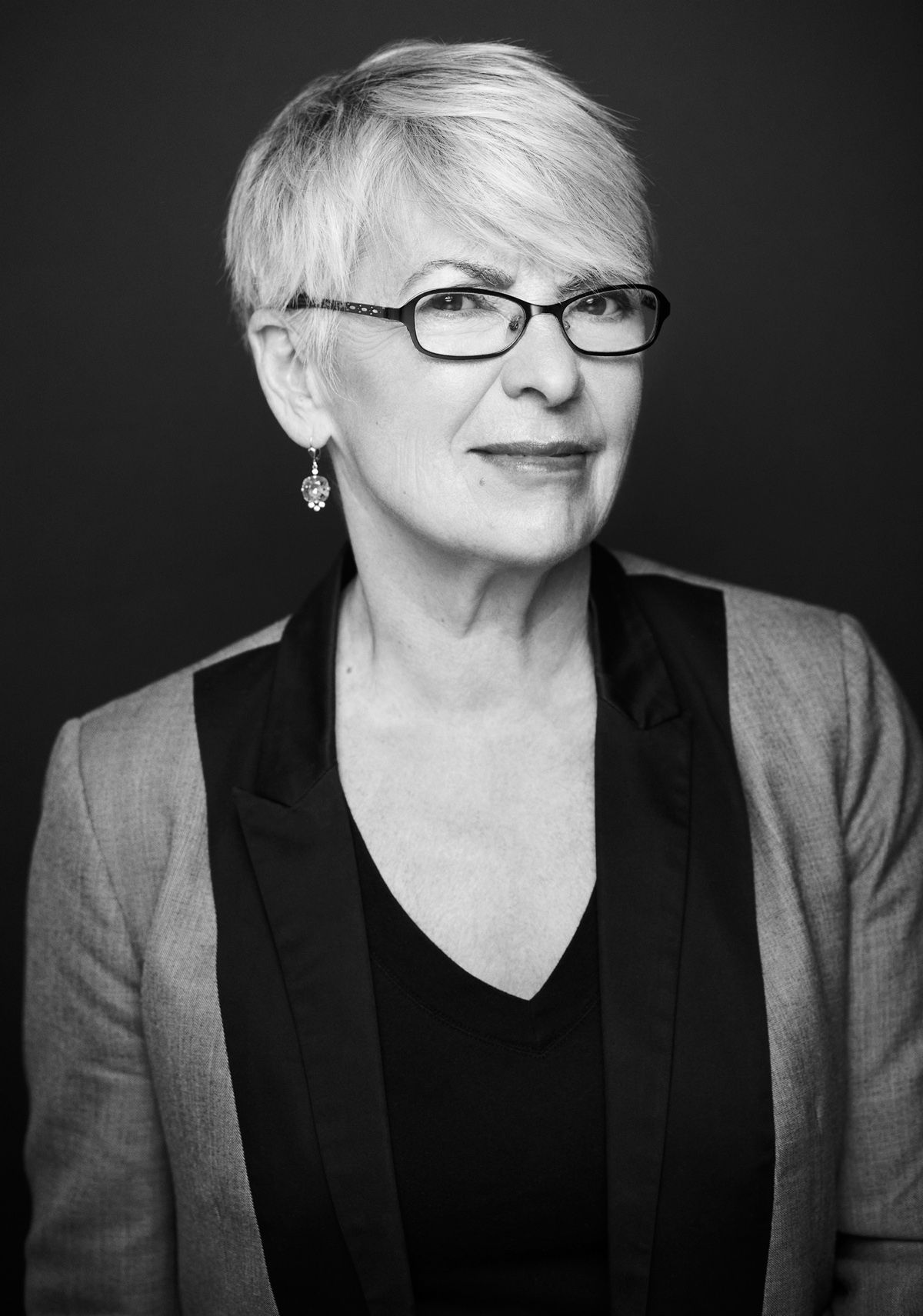 Black and white studio photograph of an older woman. She has very short gray hair, and she wears earrings, glasses, a black blouse, and a gray and black jacket. She smiles discreetly at the camera.