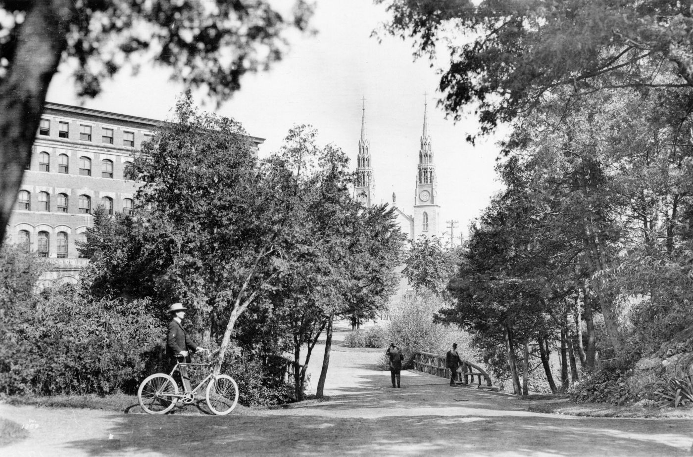 Photographie en noir et blanc d’un homme marchant à côté de sa bicyclette. Il se dirige vers une esplanade bordée d’arbres, menant à un pont de bois que traversent deux hommes à pied. Les deux clochers d’une grande église sont visibles entre les arbres.