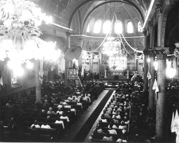 Photographie en noir et blanc de l’intérieur d’une église vue de haut. L’église est richement décorée. L’assistance est nombreuse.