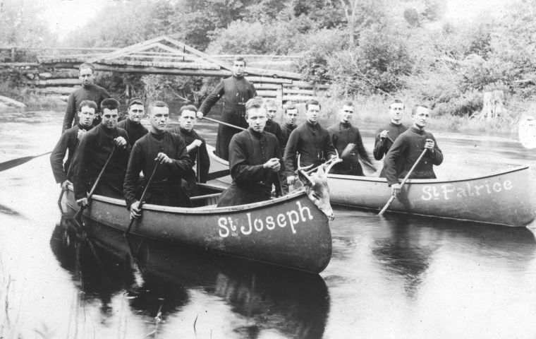 Black and white photograph of fourteen seminarians paddling on a river. They are distributed between two canoes, whose names – St Joseph and St Patrice – are painted in white on the front of the boat. The canoes are pulling away from a small wooden bridge.