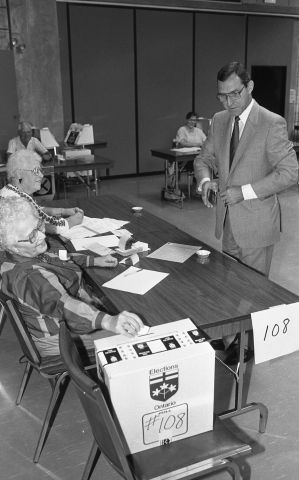 Photographie en noir et blanc d’un homme d’âge mûr en costume-cravate.  Il porte des lunettes. Il est debout devant un bureau de vote tenu par deux femmes du troisième âge. L’une d’entre elles dépose un bulletin de vote dans la boîte de scrutin. À l’arrière-plan, d’autres bureaux de vote.