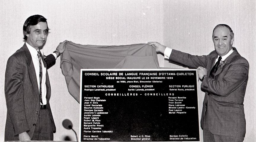 Photographie en noir et blanc de deux hommes d’âge mûr  en costume-cravate, dévoilant une plaque commémorant l’inauguration du Conseil scolaire de langue française.