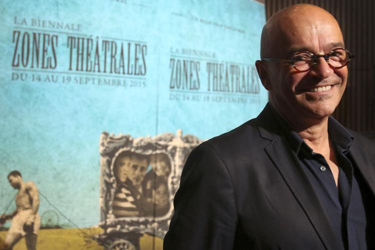 Colour photograph of a bald, smiling, middle-aged man. He wears glasses and a dark suit. He is standing in front of pale blue posters bearing the words “LA BIENNALE ZONES THÉÂTRALES DU 14 AU 19 SEPTEMBRE 2015” (THE BIANNUAL ZONES THÉÂTRALES FROM 14 TO 19 SEPTEMBER 2015.”