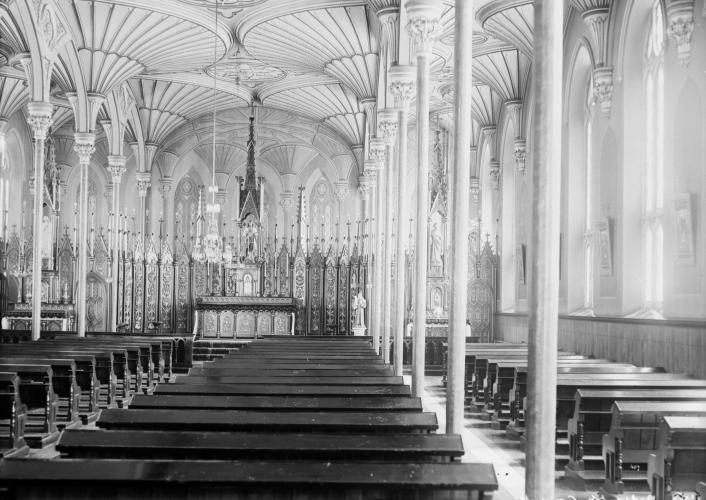 Black and white photograph of the interior of a small church, with many benches in rows. The vaulted ceiling, made of carved wood, is supported by thin columns. The altar is very ornate. orné.