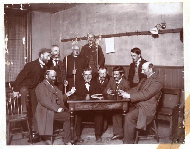 Black and white photograph of a group of men around a table. Five men are seated. Five others, including one cleric, are standing, some holding billiard cues. Two of the seated men hold cards in their hands.
