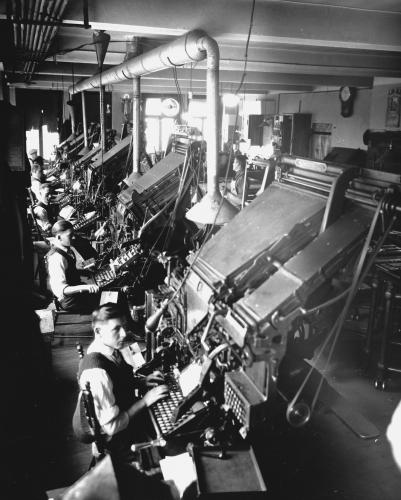 Black and white photograph of five men in shirts, vests and ties in an industrial workshop. They sit in front of imposing printing machines with keyboards.