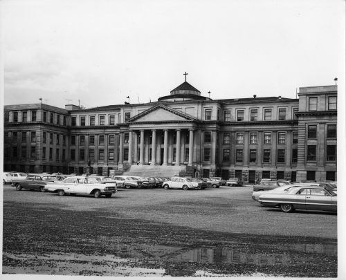 Photographie en noir et blanc d’une cinquantaine de voitures stationnées devant un imposant édifice à quatre étages construit dans le style néoclassique, avec six colonnes ioniques monumentales et un portique à fronton.