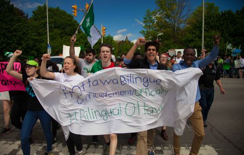 Colour photograph of five young people, fists raised, walking briskly, holding a banner with the slogan: "Ottawa bilingue officiellement/Officially bilingual Ottawa". Behind them, other protesters walk down the street waving flags and carrying placards.