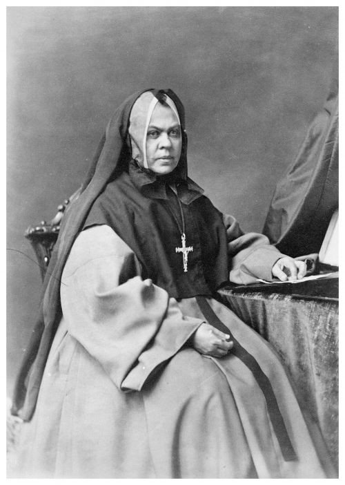 Black and white photograph of a mature nun wearing a black veil and a cross. She is sitting on a chair with a table to her left. Her expression is serious.