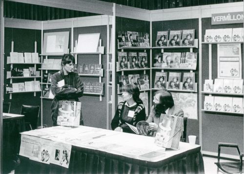 Photographie en noir et blanc d'un homme et d'une femme d'âge moyen assis à une table devant un étalage de livres. Debout à côté d'eux, une femme plus jeune. Surplombant le kiosque, un panneau avec les mots: "L'EDITION FRANCO-ONTARIENNE 461."