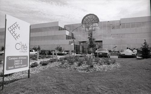 Black and white photograph of a large, three-storey building with a skylight and many windows. Cars are parked on either side of a wide entrance. A large Cité collégiale sign is positioned in the green space leading to the entrance of the building.
