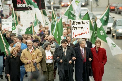 Colour photograph of a large crowd walking down a city street, demonstrating. They are carrying Franco-Ontarian flags and placards that read, “Montfort fermé : Jamais !” with the “Jamais” positioned obliquely, in stylized red letters. The six people at the front of the crowd – three men and three women, most of them older – walk with their arms linked.