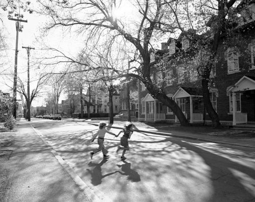 Photographie en noir et blanc de deux enfants qui traversent une rue résidentielle, main dans la main. La rue est bordée de maisons attenantes en briques, à plusieurs étages, et de grands arbres.
