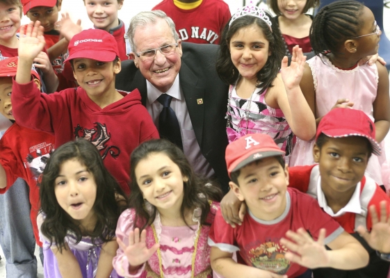 Colour photograph of an elderly man wearing a suit and tie. He sits, smiling, in the midst of a group of children representing gender and racial diversity. The children, dressed in varying shades and patterns of red and pink, also smile and wave at the camera.