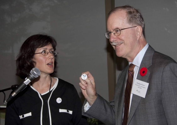 Colour photograph of two people standing in front of a microphone: a middle-aged, dark-haired women wearing a dark suit with white trim, and an older man wearing a gray suit, white shirt and brown tie. Both wear glasses. The man is holding a pin, which he is presenting to the woman.