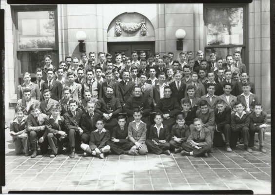 Black and white photograph of about eighty young boy, in front of a stone building. The boys in the first row are kneeling, in the next two rows sitting, and in the other five rows standing. All boys wear suits, with some also wearing ties. Three priests in cassocks are seated at the center of the group.