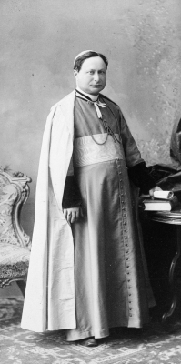 Black and white photograph of a mature man wearing the liturgical garments of an archbishop. His short brown hair is hidden under his skullcap. He stands next to a table, a document in his left hand. He suffers from a slight strabismus.