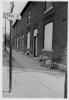 Black and white photograph of a young boy pushing a preschooler in a stroller alongside a brick building. A road sign indicates that they are at an Ottawa Street intersection.