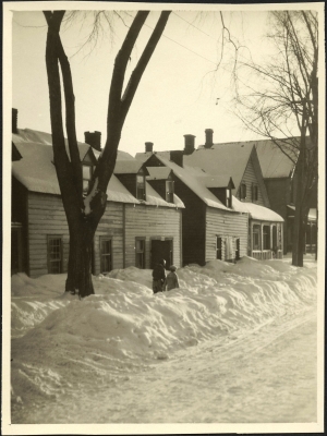 Black and white photograph of a residential street in winter, with wooden two-storey houses in need of maintenance. A couple walks along the snowy sidewalk.