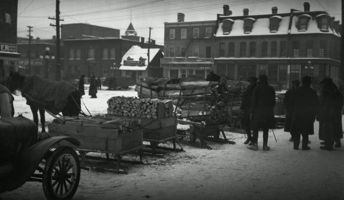 Photographie en noir en blanc d’une place de marché en hiver. Au premier plan, des traîneaux chargés de bois coupé. Plusieurs hommes vêtus de manteaux et de chapeaux devisent à côté des traîneaux.