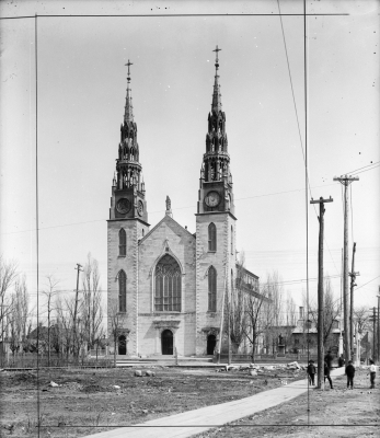 Black and white photograph of a large three-storey white stone church with a large stained glass window in the Centre front. The space in front of the church is vacant. Some small houses, as well as trees and electrical wires appear on either side of the church.
