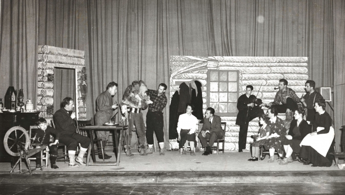 Black and white photograph of a scene from a play. About fifteen actors, men and women, dressed as early French settler and travellers. The set is the inside of a log house.