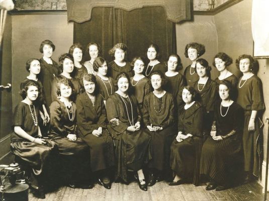 Black and white photograph depicting a group of some twenty women of various ages. Some women are seated, with others standing behind them in two rows. All wear black dresses, and many wear long pearl necklaces.