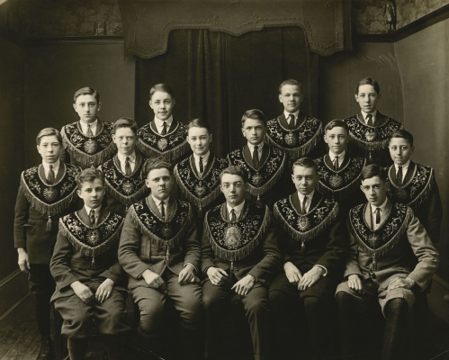 Black and white photograph depicting a group of fifteen teenage boys. Some are sitting, with others standing behind them in two rows. All wear suits and ties, as well as hoods adorned with gold embroidery and tassels.