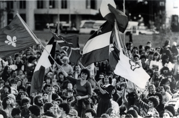 Black and white photograph of a large crowd, mostly seated. In the centre, two young women stand out from the throng, waving flags. Other people also wave flags, including two Franco-Ontarian flags and one Acadian flag.