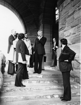 Black and white photograph of men in dark suits. They are standing under a stone arch, with some standing on a short flight of stairs leading to the arch. They hold briefcases and portfolios, with their gaze turned toward an older man standing at the top of the stairs, smoking a pipe.