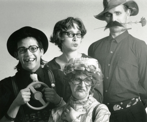 Black and white photograph of four costumed actors. One man is missing a tooth, the other wears a false mustache and holds a flower in his mouth. Between them, a women wears a cap, big glasses, and curlers; she is grimacing. The other woman is pursing her lips.