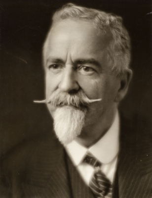 Black and white studio photograph, three-quarter view, of an elderly man with white hair, beard and mustache. He wears a white shirt, and pin-striped suit and tie, looking very distinguished.