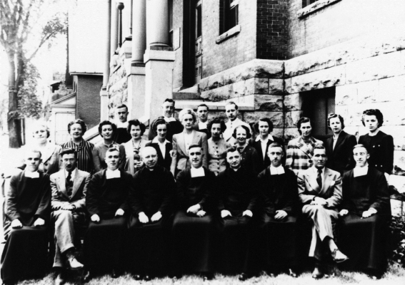Black and white photograph of a group posing in front of a stone and brick building. The group numbers around twenty people, including mostly middle-aged men, women, and a few priests wearing cassocks. Some are sitting, with others standing behind them, in two rows.