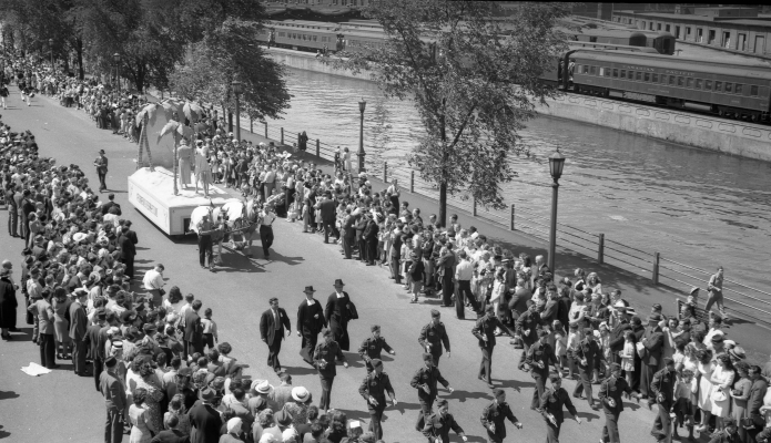 Photographie en noir et blanc d’une foule imposante, le long d’un défilé, au bord d’un canal. Devant un char allégorique tiré par des chevaux défilent des militaires et des religieux. Un train passe sur l’autre rive.