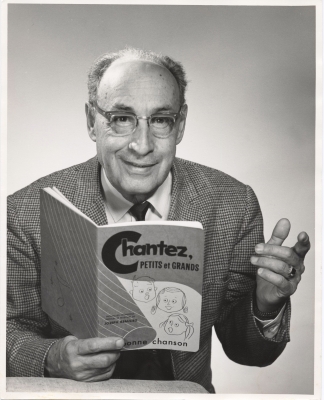 Black and white studio photograph of a mature, balding man. He wears glasses, a checked suit and a black tie. He is smiling and holding a book in his right hand, titled Chantez, petits et grands (Sing, young and old).