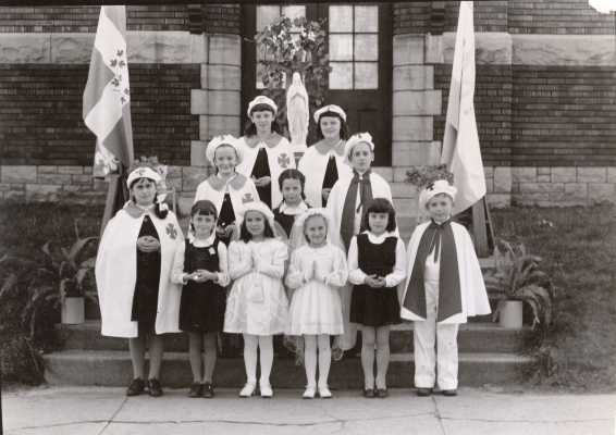 Black and white photograph showing a group of children of various ages standing in front of, or on, a staircase leading to the front door of a brick building. Some are dressed in school uniforms, others in communion dresses or special costumes with matching cape and hat.