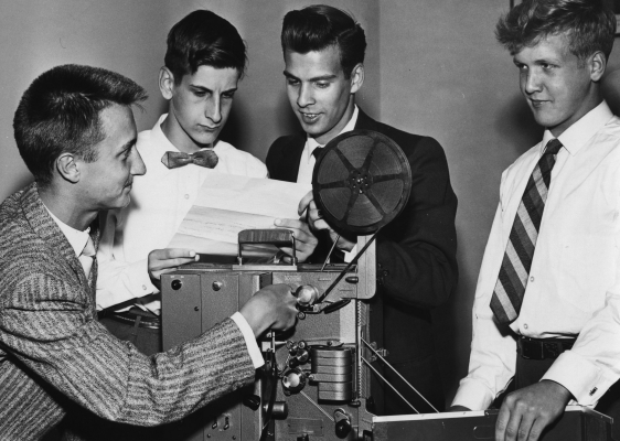 Black and white photograph of four male teens around a projector. The two young men in the center of the image are reading a document, and a third is operating the projector. They are all well-dressed.