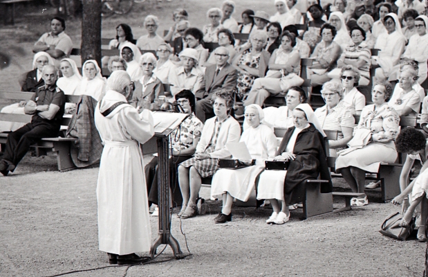 Photographie en noir et blanc d’un prêtre vu de biais, s’adressant à un auditoire, assis sur des bancs, en plein air. Il s’agit de femmes d’un certain âge pour la majorité.